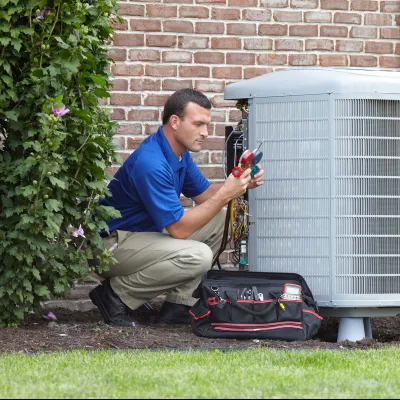 Technician in blue shirt repairing residential outdoor air conditioning unit with tools and tool bag on lawn.