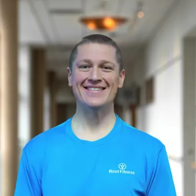Smiling man wearing a blue Reid Fitness t-shirt standing in a bright indoor hallway with windows.