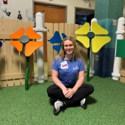Young woman in blue shirt sitting cross-legged on green floor with colorful pinwheel decorations behind her