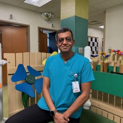 Man in teal medical scrub top sitting in a colorful indoor play area with large blue and orange flower decorations.