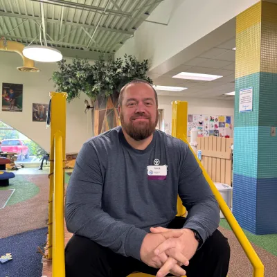 Man with a beard wearing a gray shirt sitting on yellow steps in a colorful, modern indoor space with plants and artwork.