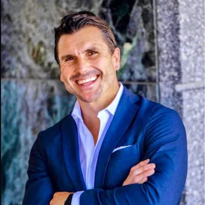 Smiling man in blue suit with arms crossed standing against a stone wall background