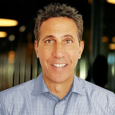 Smiling middle-aged man with short dark hair wearing a blue checkered shirt in an indoor setting with blurred lights.