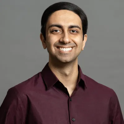Smiling man with dark hair wearing a burgundy shirt against a plain gray background.