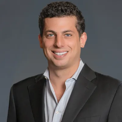 Smiling man with curly hair wearing a black blazer and checkered shirt against a plain gray background