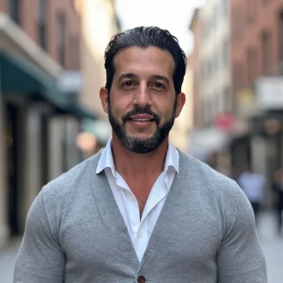 Smiling man with dark hair and beard wearing a gray cardigan and white shirt in an urban street setting.