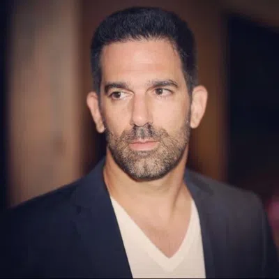 Portrait of a serious man with short dark hair and beard wearing a black blazer and white v-neck shirt