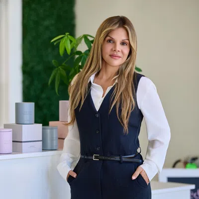 Confident woman in vest stands by table with plants and pastel-colored candles in modern interior.