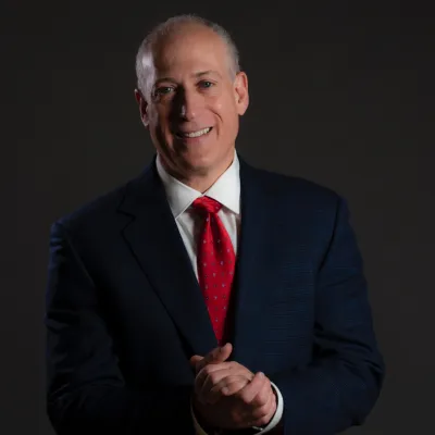 Middle-aged man in a dark suit and red tie smiling against a dark background with hands clasped.
