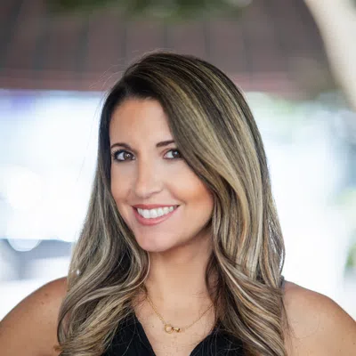 Smiling woman with long highlighted hair wearing a black sleeveless top and gold necklace, blurred background.