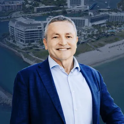 Smiling man in blue suit jacket and light blue shirt with coastal cityscape and beach in the background