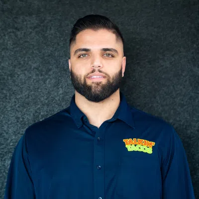 Man with beard wearing navy blue shirt with colorful Talkin' Tacos logo on black textured background