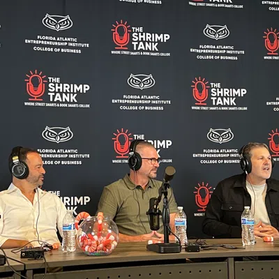 Three men wearing headphones speaking into microphones at a Shrimp Tank podcast event with branded backdrop and water bottles.