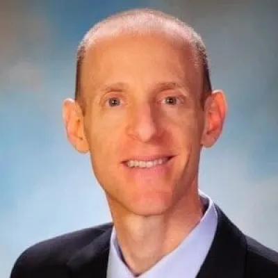 Professional headshot of a middle-aged man in a suit, light blue shirt, and red patterned tie, smiling against a soft blue background