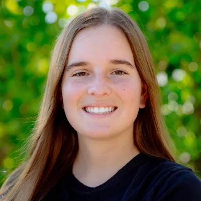 Smiling young woman with long brown hair wearing a black shirt standing outdoors with green foliage background