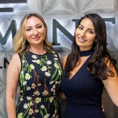 Two women smiling, one in a floral dress and the other in a navy blue dress, standing against a textured wall.