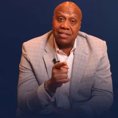 Man in light gray checkered blazer and white shirt pointing forward against a dark blue background.