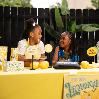 Two smiling girls at a bright lemonade stand with lemons, cups, and fresh lemonade outdoors.