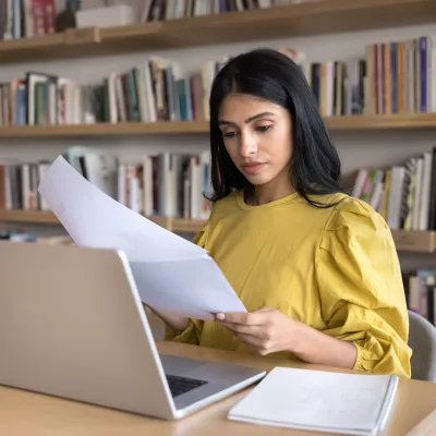 Young woman in a yellow blouse reviewing papers while working on a laptop in a library setting.