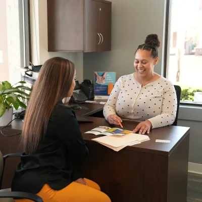 Two women discussing documents across a desk in a bright office with a potted plant and window light.