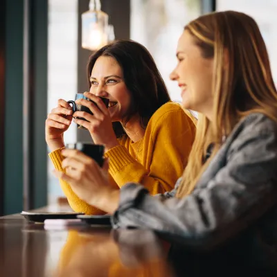 Two women enjoying coffee and smiling while sitting by the window in a cozy cafe.