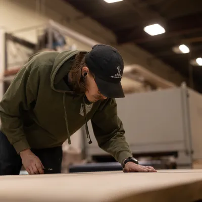 Young man in green hoodie and black cap carefully measuring wood in a workshop environment.