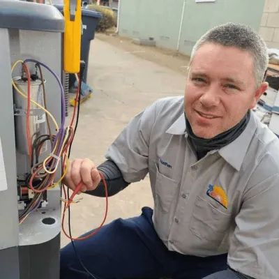 Technician in uniform testing HVAC system wiring outdoors with multimeter, smiling at camera