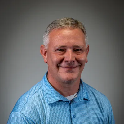 Middle-aged man with short gray hair wearing a light blue polo shirt smiling against a gray background