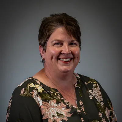Smiling woman with short brown hair wearing a dark floral blouse against a grey background