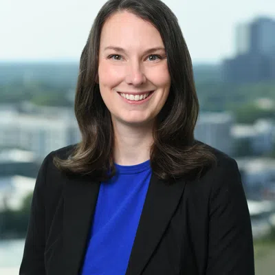 Smiling woman with dark hair wearing a blue top and black blazer in an office with cityscape background.