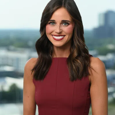 Smiling woman with long brown hair wearing a sleeveless burgundy dress with cityscape background.
