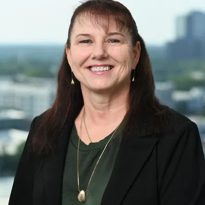 Smiling middle-aged woman with long brown hair wearing black blazer and green top against cityscape background