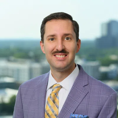 Professional man with mustache wearing a purple checkered suit and yellow striped tie standing outdoors.