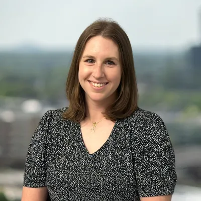 Smiling woman with shoulder-length brown hair wearing a black patterned blouse standing outdoors with blurred city background.