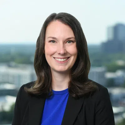 Smiling woman with dark hair wearing a blue top and black blazer in an office with cityscape background.