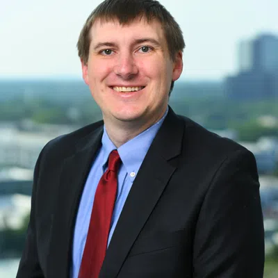 Smiling man in black suit, blue shirt, and red tie posing outdoors with blurred cityscape background.