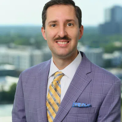 Professional man with mustache wearing a purple checkered suit and yellow striped tie standing outdoors.
