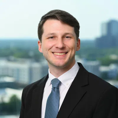 Smiling young man in black suit and blue tie standing outdoors with blurred cityscape in background
