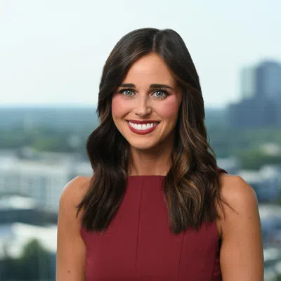 Smiling woman with long brown hair wearing a sleeveless burgundy dress with cityscape background.