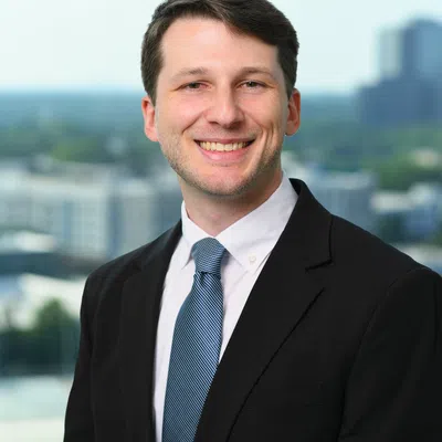 Smiling young man in black suit and blue tie standing outdoors with blurred cityscape in background