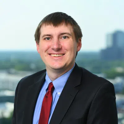 Smiling man in black suit, blue shirt, and red tie posing outdoors with blurred cityscape background.