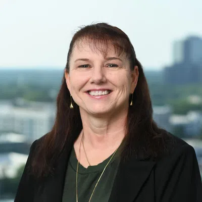 Smiling middle-aged woman with long brown hair wearing black blazer and green top against cityscape background