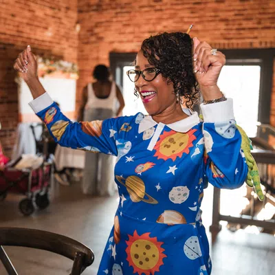 Smiling woman in colorful space-themed dress raises hands in a bright, cozy room with brick walls and wooden furniture.