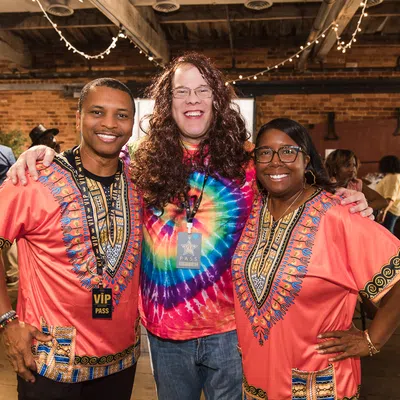 Three smiling people in vibrant attire pose inside a warmly lit event space with brick walls and string lights.