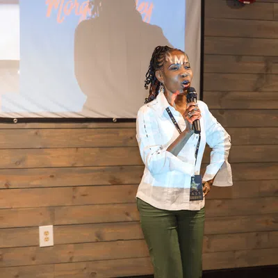 Woman speaking with microphone in front of a wood-paneled wall and projector screen with overlaid text on it.