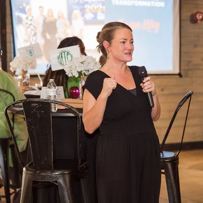 Woman in black dress speaking into a microphone at an indoor event with seated guests and wooden walls.