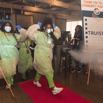 Healthcare workers in protective gear walking on a red carpet, with overlaid text on signs showing VIP and community partner logos.