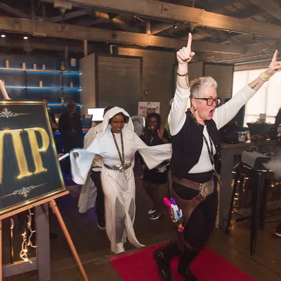 Person excitedly raising arms near VIP sign with costumed individuals in a lively indoor event space.