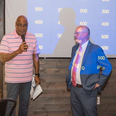Two men standing in front of a score board projection in a casual indoor setting during an event or game night.