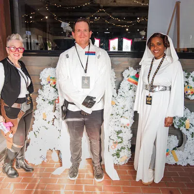 Three people in themed costumes standing in front of a white floral display spelling out 'ABS' inside a venue.